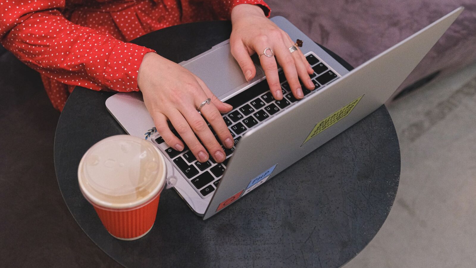 Een vrouw in een rode stippen jurk typt op een laptop met koffie in de buurt.