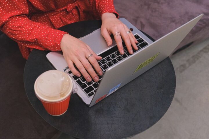 Een vrouw in een rode stippen jurk typt op een laptop met koffie in de buurt.