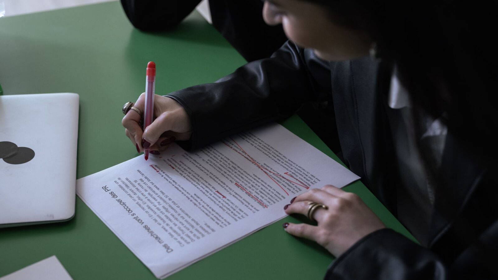 Een vrouw in een kostuum gebruikt een rode pen om een afgedrukt document te bewerken op een groene tafel met een laptop.
