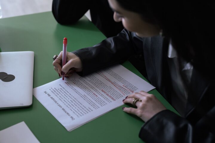 Een vrouw in een kostuum gebruikt een rode pen om een afgedrukt document te bewerken op een groene tafel met een laptop.